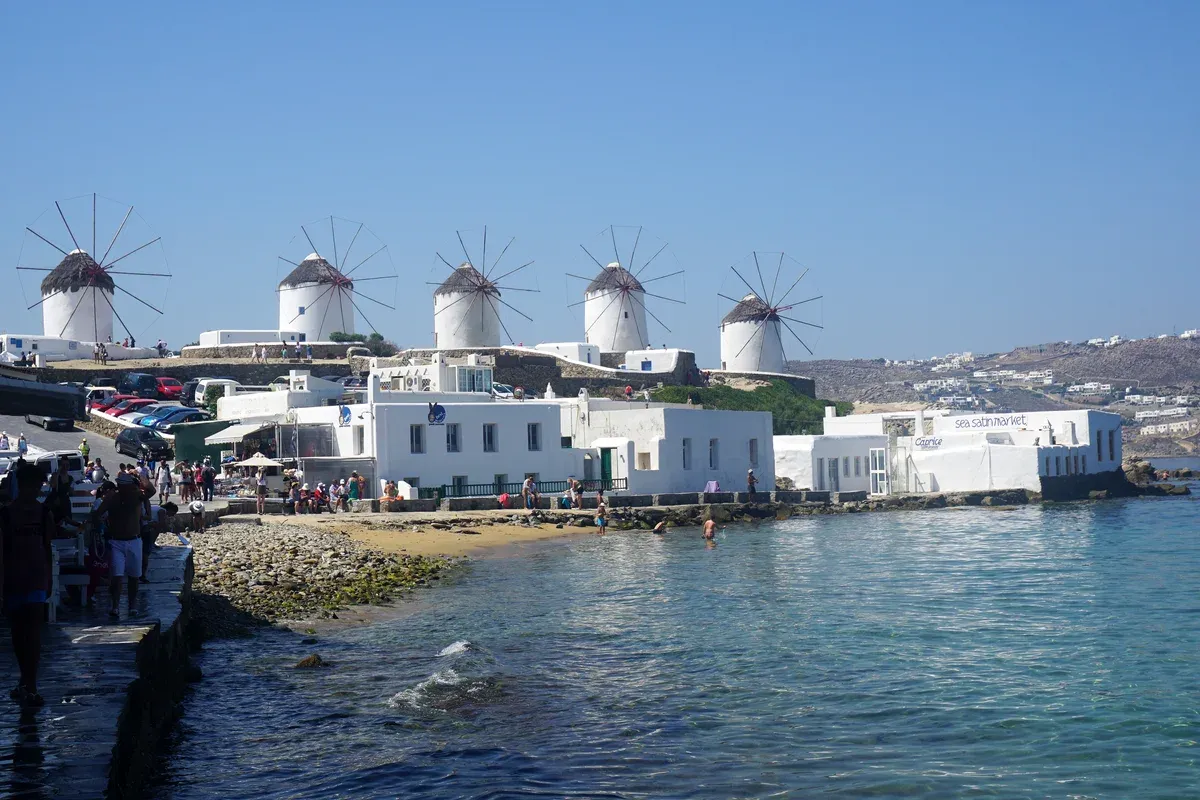 Crowded waterfront below the Kato Mili windmills in harsh midday June light, the slot that weakens a Mykonos photoshoot
